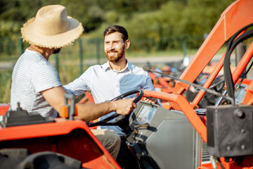 Young agronomist with elegant salesman choosing a tractor for farming on the open ground of...