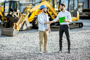 Builder choosing heavy machinery for construction with a sales consultant shaking hands on the open...