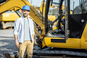 Portrait of a handsome builder standing near the escavator on the open ground of the shop with heavy machinery