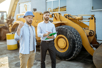 Builder choosing heavy machinery for construction with a sales consultant, signing some documents on the open ground of a shop with special vehicles