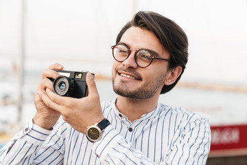 Photo of cheerful caucasian man taking photo on retro camera and smiling while walking on pier