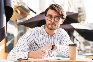 Photo of confident unshaven man wearing eyeglasses writing on diary and drinking coffee