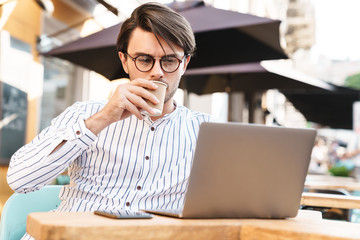 Fototapeta premium Photo of brooding caucasian using laptop and cellphone while drinking coffee in cafe outdoors