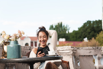 Portrait of joyous brunette woman holding smartphone and drinking coffee while resting on terrace outdoors