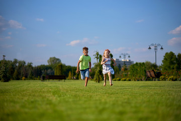 Fototapeta premium Two happy little kids having fun while running through the grassy field and racing against each other. children running around the green lawn, happy and cheerful, running away to the summer park