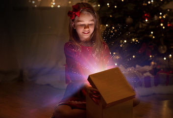 christmas, holidays and childhood concept - happy little girl opening gift box with magic light in the dark at home