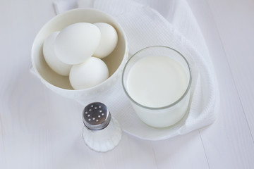 four white eggs lie in a plate on a white wooden table next to a towel and a glass of milk and a salt shaker.