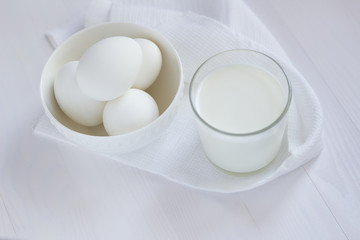 Fototapeta na wymiar four white eggs lie in a plate on a white wooden table next to a towel and a glass of milk.