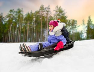 childhood, sledging and season concept - happy little kids sliding on sled down snow hill in winter over forest background