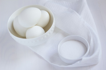 four white eggs lie in a plate on a white wooden table next to a towel and salt shaker.
