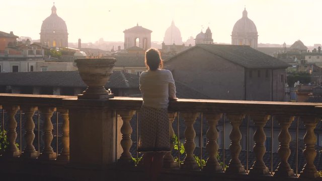 Young woman tourist in fashion white dress with hat walking at panoramic view of Rome cityscape from campidoglio terrace at sunset. Landmarks, domes of Rome, Italy.