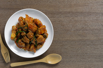 top view of stir fried pork belly with red curry paste in a ceramic dish on wooden table. hot and spicy thai style food menu.