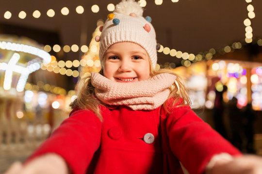 holidays, childhood and people concept - happy little girl at christmas market taking selfie in winter evening