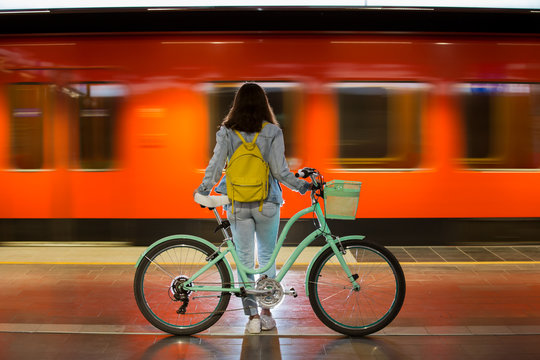 Teenager girl in jeans with yellow backpack and bike standing on metro station, waiting for train, smiling, laughing. Orange train passing by behind the girl. Futuristic subway station. Finland, Espoo