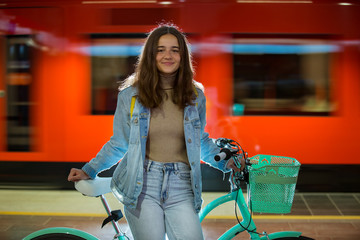 Teenager girl in jeans with yellow backpack and bike standing on metro station, waiting for train, smiling, laughing. Orange train passing by behind the girl. Futuristic subway station. Finland, Espoo