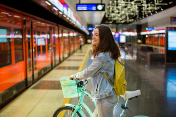 Teenager girl in jeans with yellow backpack and bike standing on metro station, waiting for train,...