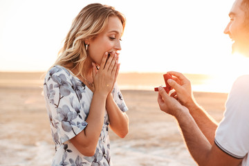 Photo of smiling man making proposal to his amazed woman with ring while walking on sunny beach - Powered by Adobe