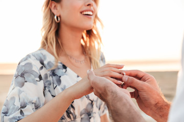 Cropped photo of young man putting engagement ring on woman's finger while walking on beach