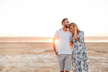 Photo of joyful young couple smiling and hugging while walking on sunny beach - Powered by Adobe