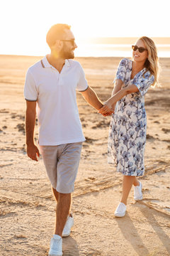 Photo of happy young couple smiling and holding hands together while walking on sunny beach