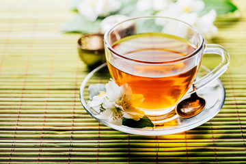 Glass of hot herbal tea on bamboo background closeup. Teacup with organic green tea and fresh jasmine flower on wooden table.