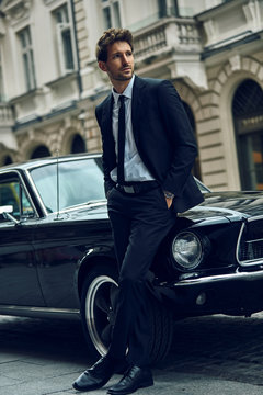 Young handsome man with black classic car wearing black suit with hands in pocket