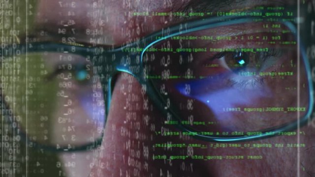 Male hacker working on a computer for cyber attack while green binary hacking code characters reflect on his face in a dark office room