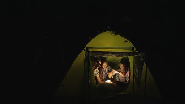 Family with a dog in a camping tent at night reading a book, smiling and playing, doing a spell over a book.