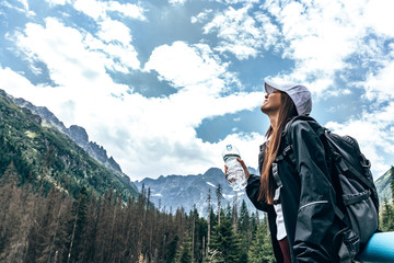 Young beautiful girl traveler in white hat and black jacket and backpack, standing with closed eyes and holding a bottle of water, amazing mountains on the background
