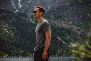 Naklejka premium Side view portrait of young man traveler in black sunglasses and grey t-shirt in front of the mountain lake and range, active life concept
