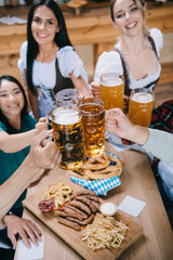 selective focus of attractive waitresses in traditional german costumes clinking mugs of beer with multicultural friends