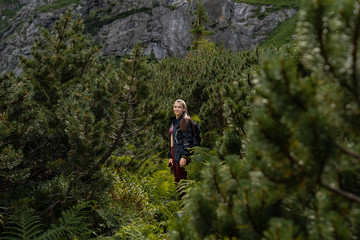 Naklejka na ściany i meble Young girl traveler in balck jacket standing among coniferous forest in the mountains, active lifestyle concept