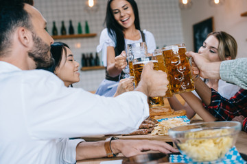 selective focus of waitress in traditional german costume holding glass of beer while standing near multicultural friends celebrating octoberfest in pub