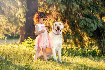Naklejka na ściany i meble Cute adorable little curly Caucasian girl wearing yellow sunglasses with her dog in park outside at sunset on summer day. Child playing with animal domestic pet. Happy childhood concept.