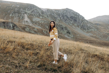 Naklejka premium Stylish girl in rustic dress smiling and waving hair in sunny meadow in mountains. Portrait of happy boho woman in countryside at sunset, positive vibes, rural simple life. Atmospheric image