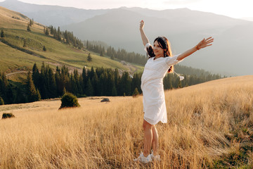 Fototapeta premium Stylish girl in rustic dress smiling and waving hair in sunny meadow in mountains. Portrait of happy boho woman in countryside at sunset, positive vibes, rural simple life. Atmospheric image