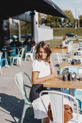young woman in a white blouse and skirt sits on a summer terrace of a cafe in the city on a sunny morning. Stylish lifestyle