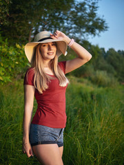 Outdoor lifestyle young happy blonde woman wearing trendy straw hat dressed red blouse blue denim jeans shorts emotionally posing green meadow in evening warm autumn sun