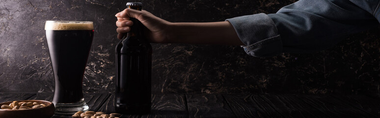 cropped view of man holding bottle near glass of dark beer and bowls with nuts on wooden table