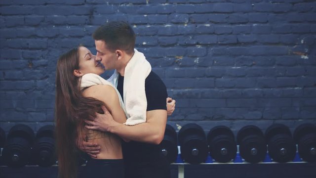 Young sporty couple guy and girl are in the gym. After a training session, they embrace themselves kissing and looking at each other in the eye. During this, they have white towels.