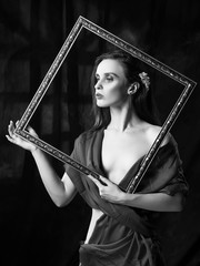 Beautiful girl with trendy makeup, wearing a silk scarf thrown over her shoulders that covers her naked breast, holds picture frame on the background of the shadow from the window. Black and white