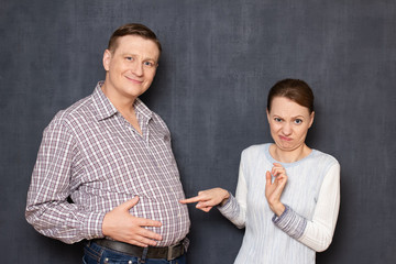 This is disgusting! Studio waist-up shot of dissatisfied woman touching with disgust and displeasure big stomach of happy satisfied smiling man standing nearby over gray background. Man's obesity