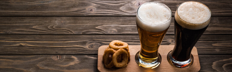 panoramic shot of glasses of dark and light beer with foam near fried onion rings on wooden table