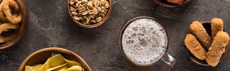 panoramic shot of mug of light beer near peanuts and crisps on brown textured surface