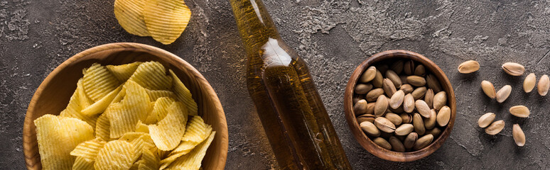 panoramic shot of bottle of light beer near bowls with pistachios and crisps on brown textured surface
