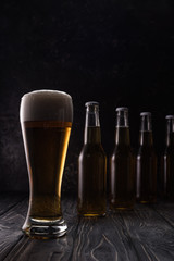 glass of fresh light beer with foam near bottles on wooden table