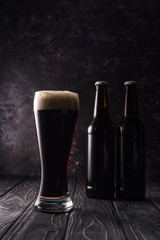 glass of beer with foam near bottles on wooden table