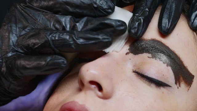 A permanent master in black gloves is slowly wiping with a white napkin brown pigment from the eyebrow of a lady client after the microblading procedure, close-up.