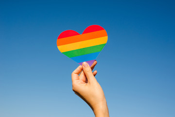 Woman holds in hands a heart in the colors of the rainbow. Young beautiful girl. LGBT history month. Pride Month. Lesbian Gay Bisexual Transgender. LGBT flag. Love, human rights, tolerance. LGBTQ+ - obrazy, fototapety, plakaty