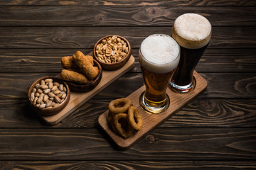 glasses of dark and light beer near bowls with peanuts, pistachios, fried cheese and onion rings on wooden table